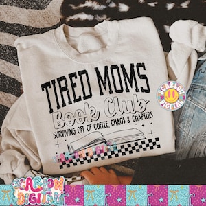 Op de afbeelding: Een crèmekleurige sweatshirt met de tekst "TIRED MOMS Book Club" in het zwart. Daaronder staat "SURVIVING OFF OF COFFEE, CHAOS & CHAPTERS" en een illustratie van een open boek. Het ontwerp bevat een zwart-wit geblokt patroon.