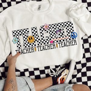 May include: White sweatshirt with the word "TEACHER" in black and white checkered letters. The letters are decorated with colorful flowers, a rainbow, a peace sign, a smiley face, and a pencil.