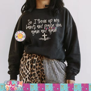 Op de afbeelding: Zwart sweatshirt met de tekst "So I throw up my hands and praise you again and again." Het ontwerp bevat een kruis over een open boek. Een kleurrijk logo staat aan de linkerkant van het sweatshirt.