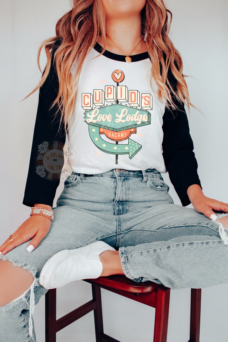 以下が含まれることがあります： 白と黒のベースボールTシャツに、レトロなデザインの看板がプリントされています。看板には「Cupid's Love Lodge Vacant」と書いてあります。看板は、ターコイズブルー、ピンク、オレンジの色合いで、白い水玉模様が描かれています。このTシャツを着ているのは、長いブロンドの髪をした人です。