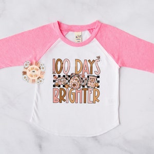 May include: A pink and white long-sleeved shirt with a  "100 Days Brighter" graphic. The graphic features a checkered background with school supplies and flowers.