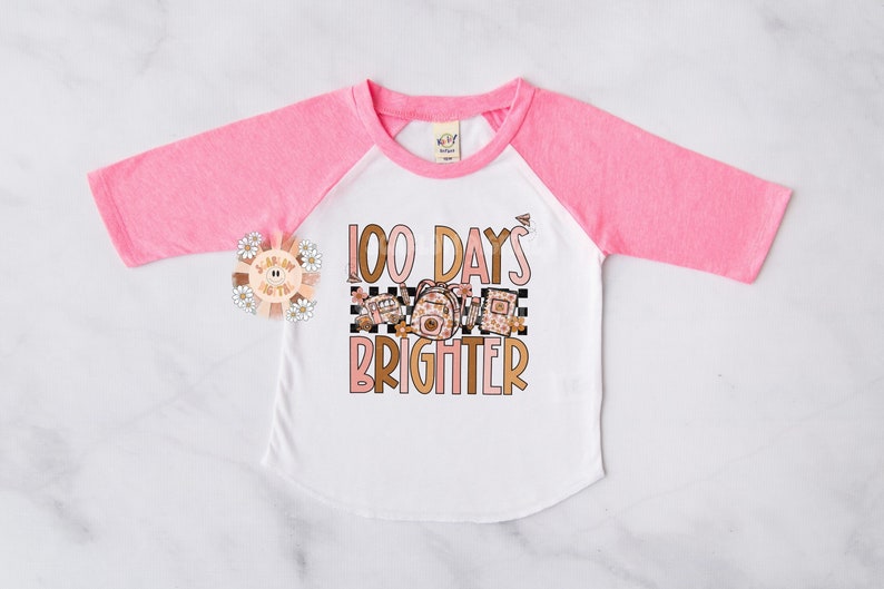 May include: A pink and white long-sleeved shirt with a  "100 Days Brighter" graphic. The graphic features a checkered background with school supplies and flowers.