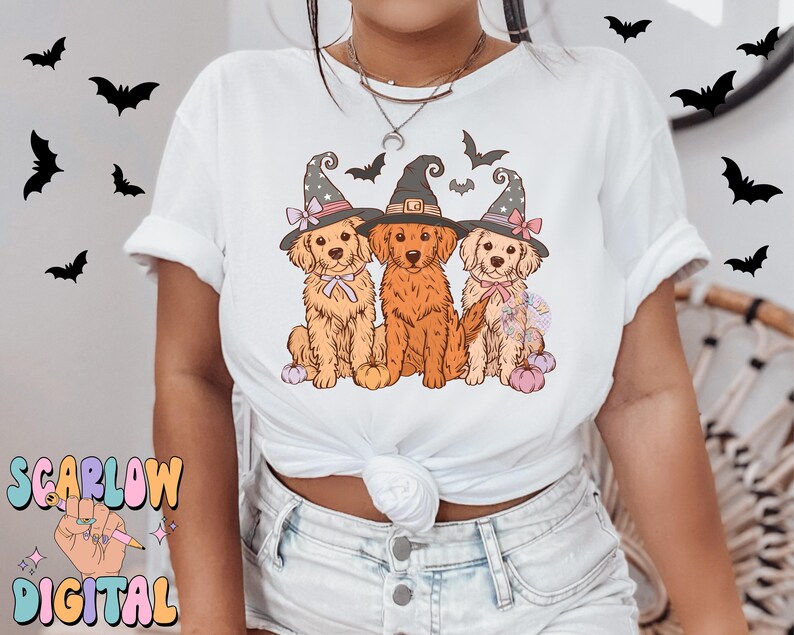 K&ouml;nnte beinhalten: Wei&szlig;es T-Shirt mit einem Motiv von drei Golden Retrievern, die Hexenh&uuml;te tragen und K&uuml;rbisse halten. Die Hunde tragen rosa und wei&szlig;e Schleifen.