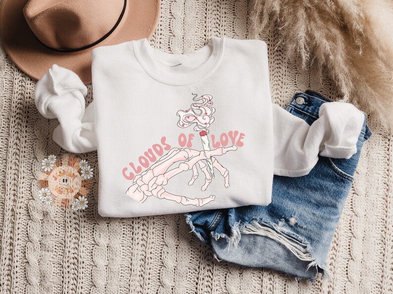 May include: White sweatshirt with a graphic design featuring a skeleton hand holding a lit cigarette with smoke that reads "Clouds of Love" in pink. The sweatshirt is paired with denim shorts.