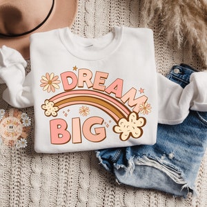 May include: A white sweatshirt with a pink and brown rainbow graphic and the text "DREAM BIG" in pink letters. The rainbow is decorated with small yellow flowers.