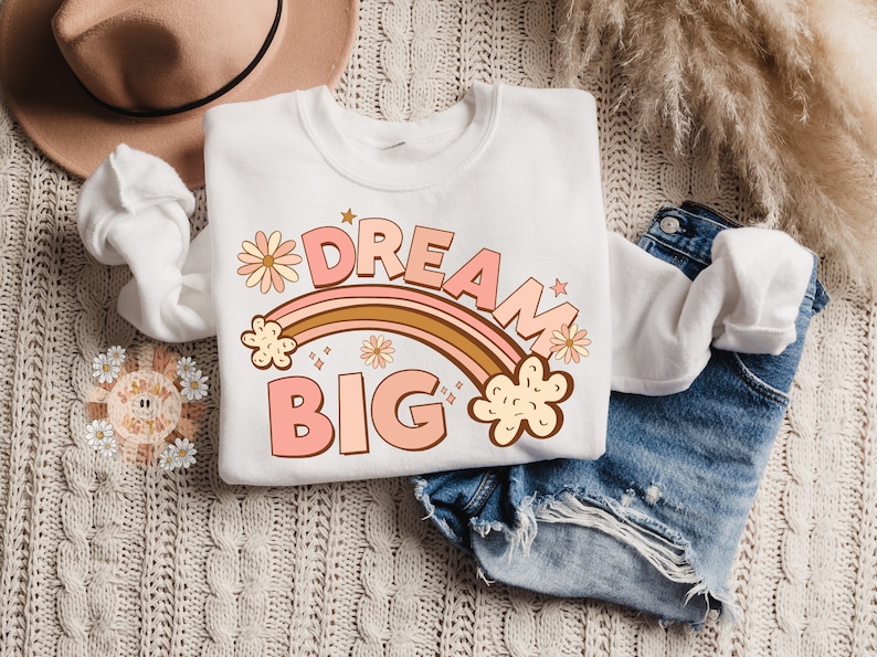 May include: A white sweatshirt with a pink and brown rainbow graphic and the text "DREAM BIG" in pink letters. The rainbow is decorated with small yellow flowers.