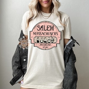 Pu&ograve; includere: Una maglietta bianca con un design grafico rosa e bianco che dice "Salem Massachusetts Apothecary Est. 1692". La grafica presenta un'illustrazione in stile vintage di cinque bottiglie di pozioni.