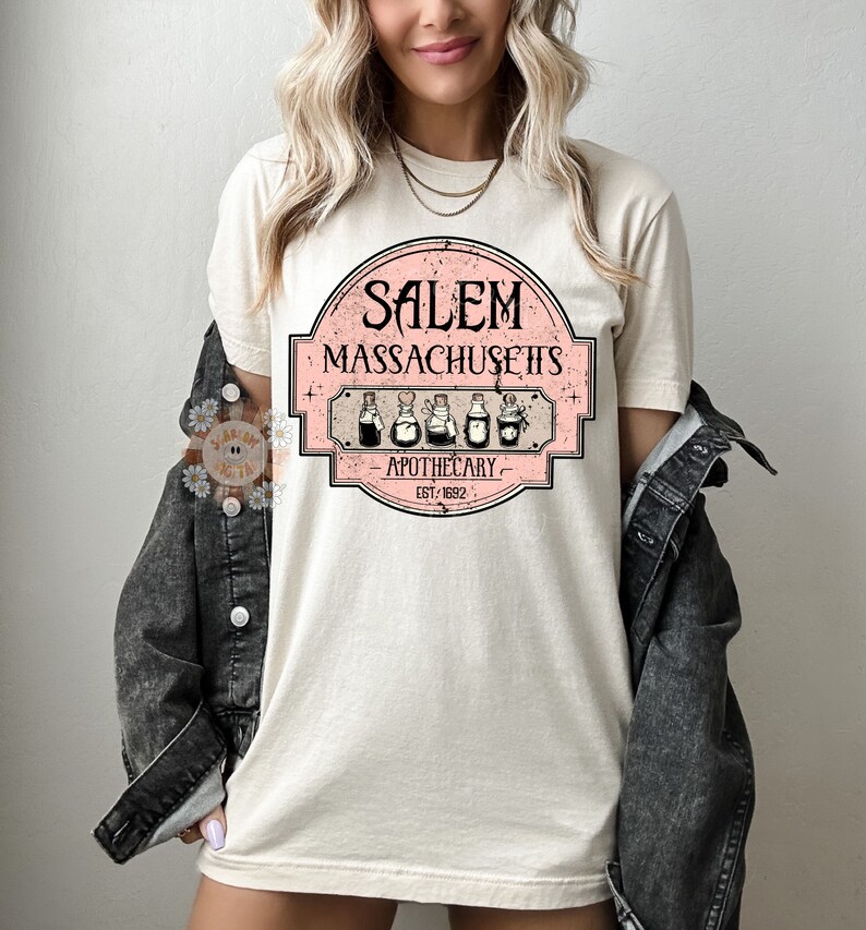 Pu&ograve; includere: Una maglietta bianca con un design grafico rosa e bianco che dice "Salem Massachusetts Apothecary Est. 1692". La grafica presenta un'illustrazione in stile vintage di cinque bottiglie di pozioni.