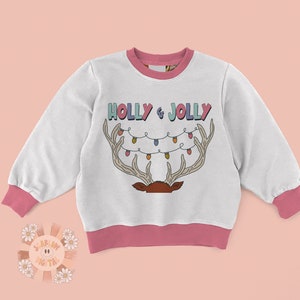 Puede incluir: Sudadera blanca con ribete rosa y un dise&ntilde;o navide&ntilde;o. El dise&ntilde;o presenta un reno con cuernos y una cadena de luces que dice "HOLLY & JOLLY".