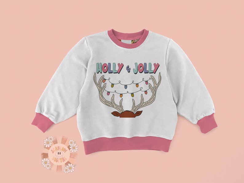 Puede incluir: Sudadera blanca con ribete rosa y un dise&ntilde;o navide&ntilde;o. El dise&ntilde;o presenta un reno con cuernos y una cadena de luces que dice "HOLLY & JOLLY".