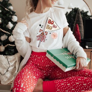 May include: A white t-shirt with a red, white, and gold graphic of a Christmas pennant with the text "Ho Ho Ho" and a candy cane. The person is wearing red and white polka dot pajamas and holding a green gift box with the text "Tis the Season".