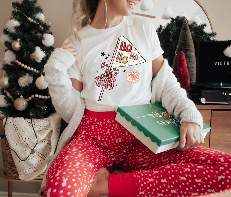 May include: A white t-shirt with a red, white, and gold graphic of a Christmas pennant with the text "Ho Ho Ho" and a candy cane. The person is wearing red and white polka dot pajamas and holding a green gift box with the text "Tis the Season".