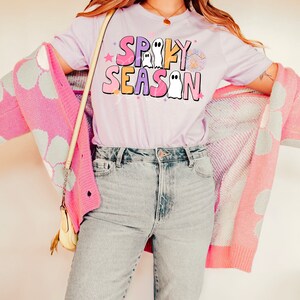 Op de afbeelding: Een lichtpaarse T-shirt met de tekst "Spooky Season" in kleurrijke letters met spookillustraties.