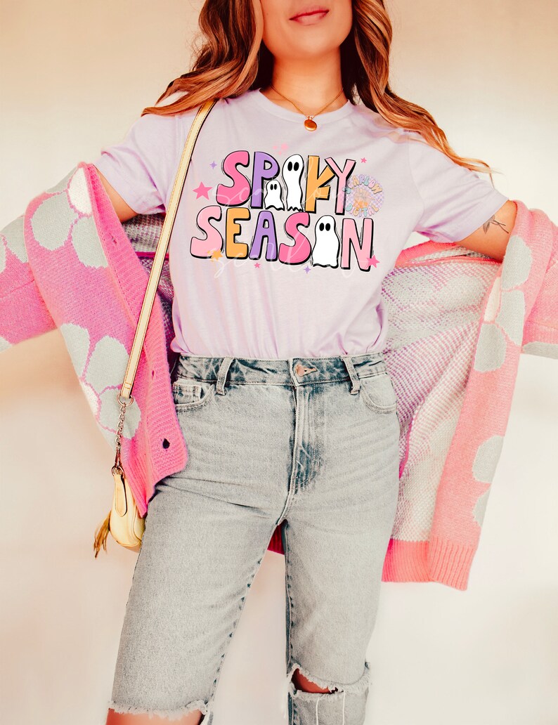 Op de afbeelding: Een lichtpaarse T-shirt met de tekst "Spooky Season" in kleurrijke letters met spookillustraties.