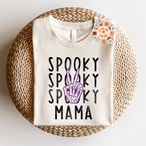 K&ouml;nnte beinhalten: Ein wei&szlig;es T-Shirt mit dem Text "Spooky Mama" in schwarzen Buchstaben. Eine violette Skeletthand mit einem Peace-Zeichen befindet sich in der Mitte des Textes.