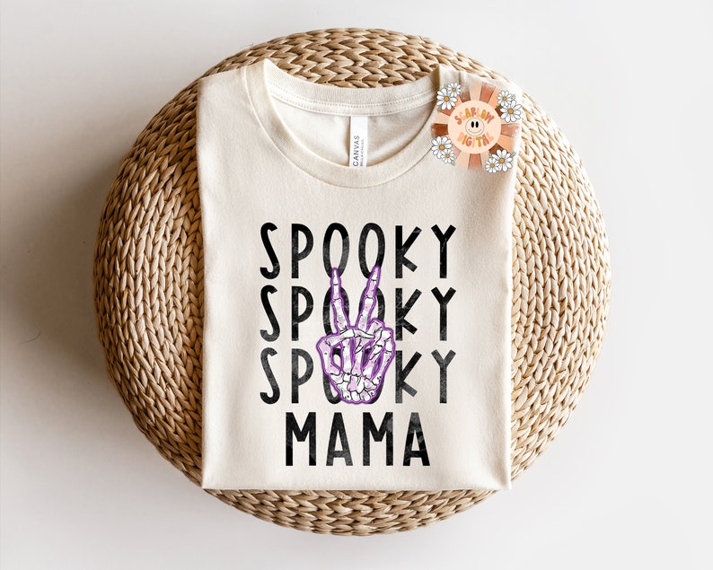 K&ouml;nnte beinhalten: Ein wei&szlig;es T-Shirt mit dem Text "Spooky Mama" in schwarzen Buchstaben. Eine violette Skeletthand mit einem Peace-Zeichen befindet sich in der Mitte des Textes.