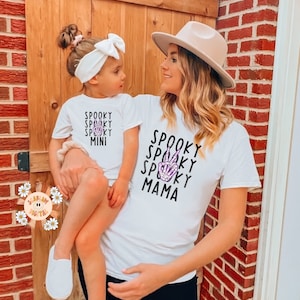 K&ouml;nnte beinhalten: Eine Mutter und ihre Tochter tragen passende wei&szlig;e T-Shirts mit einem gruseligen Halloween-Design. Das Shirt der Mutter tr&auml;gt den Schriftzug "Spooky Mama" mit einem violetten Skelett-Hand-Friedenszeichen. Das Shirt der Tochter tr&auml;gt den Schriftzug "Spooky Mini".