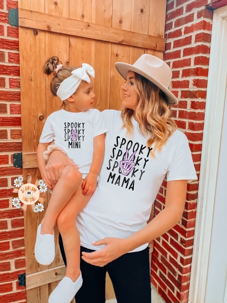 K&ouml;nnte beinhalten: Eine Mutter und ihre Tochter tragen passende wei&szlig;e T-Shirts mit einem gruseligen Halloween-Design. Das Shirt der Mutter tr&auml;gt den Schriftzug "Spooky Mama" mit einem violetten Skelett-Hand-Friedenszeichen. Das Shirt der Tochter tr&auml;gt den Schriftzug "Spooky Mini".