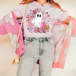 Pu&ograve; includere: Una maglietta lilla chiaro con una grafica di un fantasma che tiene una zucca e un lecca-lecca. La grafica &egrave; circondata da fiori e stelle. Il testo "Spooky Season" &egrave; scritto in lettere rosa e bianche.