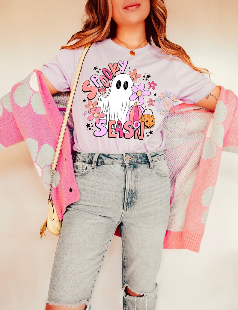 Pu&ograve; includere: Una maglietta lilla chiaro con una grafica di un fantasma che tiene una zucca e un lecca-lecca. La grafica &egrave; circondata da fiori e stelle. Il testo "Spooky Season" &egrave; scritto in lettere rosa e bianche.