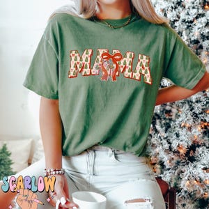 May include: A green t-shirt with the word "MAMA" printed on it in red and white letters. The letters are decorated with a pattern of Christmas trees and bows.