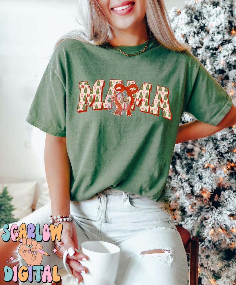 May include: A green t-shirt with the word "MAMA" printed on it in red and white letters. The letters are decorated with a pattern of Christmas trees and bows.