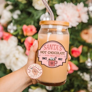 Op de afbeelding: Een glazen pot met een houten deksel en een roze en witte sticker met de tekst "Santa's Hot Chocolate - Made Fresh Daily - Since 1862". De pot is gevuld met een lichtbruine vloeistof en heeft een doorzichtige glazen rietje die uit de bovenkant steekt.
