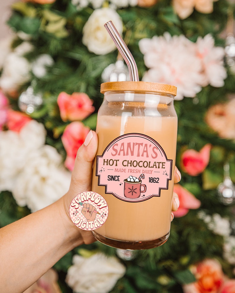 Op de afbeelding: Een glazen pot met een houten deksel en een roze en witte sticker met de tekst "Santa's Hot Chocolate - Made Fresh Daily - Since 1862". De pot is gevuld met een lichtbruine vloeistof en heeft een doorzichtige glazen rietje die uit de bovenkant steekt.