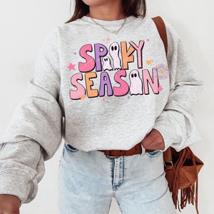 Op de afbeelding: Een grijs sweatshirt met een kleurrijke afbeelding met de tekst "Spooky Season" met geesten en sterren. Het sweatshirt wordt gedragen door een persoon met een bruine leren tas.
