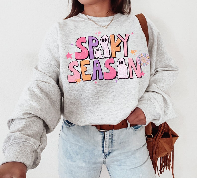 Op de afbeelding: Een grijs sweatshirt met een kleurrijke afbeelding met de tekst "Spooky Season" met geesten en sterren. Het sweatshirt wordt gedragen door een persoon met een bruine leren tas.