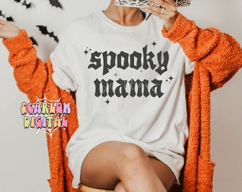 May include: A white t-shirt with the text "spooky mama" in black gothic font. The shirt is decorated with small black stars. The model is wearing the shirt with an orange cardigan and black and orange socks with a pumpkin design.