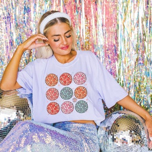 May include: A woman wearing a white headband and a lavender t-shirt with a colorful disco ball pattern. She is sitting on a sequined skirt and surrounded by silver disco balls.