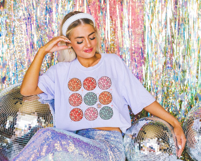 May include: A woman wearing a white headband and a lavender t-shirt with a colorful disco ball pattern. She is sitting on a sequined skirt and surrounded by silver disco balls.