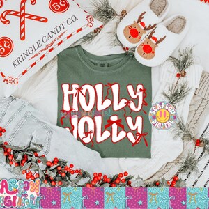 以下が含まれることがあります： お祝いのテーマのフラットレイ。白い文字と赤いリボンで「HOLLY JOLLY」と書かれた緑色のTシャツは、キャンディケインの枕、トナカイのスリッパ、赤いベリーで飾られたデニムジーンズに囲まれています。季節感のある居心地の良いシーンです。