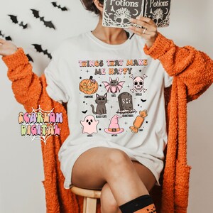 Puede incluir: Camiseta blanca con un gr&aacute;fico de iconos de Halloween y el texto "Things that make me happy". Los iconos incluyen calabazas, murci&eacute;lagos, calaveras, gatos negros, fantasmas, brujas, cementerios y caramelos.