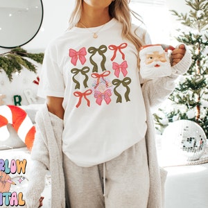 May include: A white t-shirt with a festive design featuring multiple pink and green bows. The bows are arranged in a pattern on the front of the shirt.