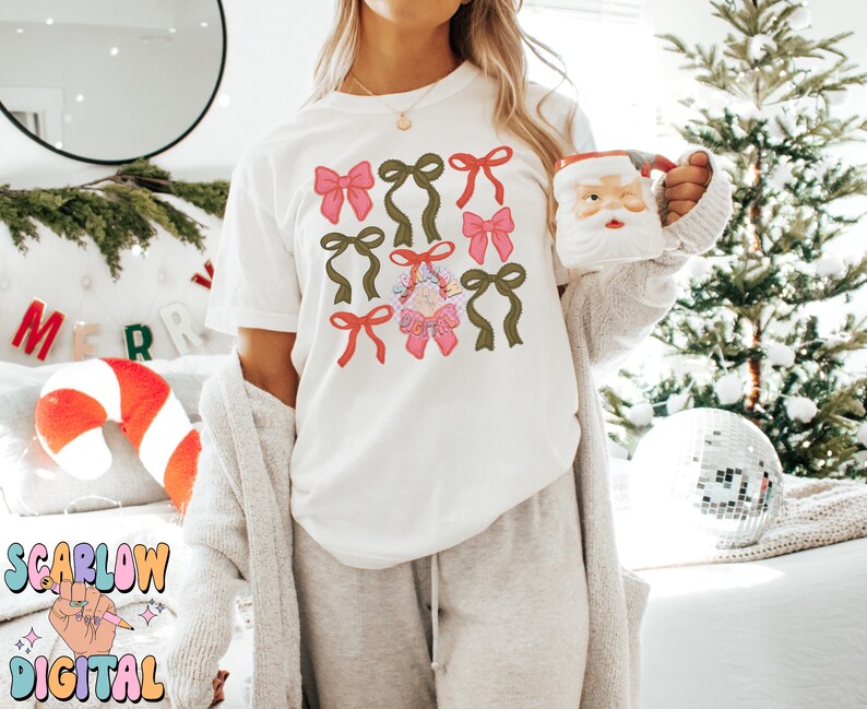 May include: A white t-shirt with a festive design featuring multiple pink and green bows. The bows are arranged in a pattern on the front of the shirt.
