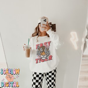 Puede incluir: Sudadera blanca con un gr&aacute;fico de un tigre con el texto "Easy Tiger" en rojo. La modelo lleva pantalones a cuadros blanco y negro y una gorra de b&eacute;isbol blanca.