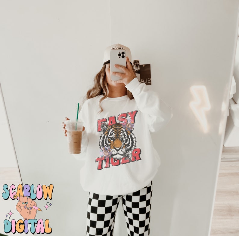 Puede incluir: Sudadera blanca con un gr&aacute;fico de un tigre con el texto "Easy Tiger" en rojo. La modelo lleva pantalones a cuadros blanco y negro y una gorra de b&eacute;isbol blanca.