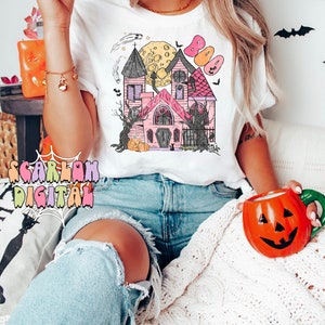 May include: A white t-shirt with a pink and white illustration of a haunted house with a full moon and bats. The text "BOO" is above the house. The model is wearing the shirt and blue jeans.