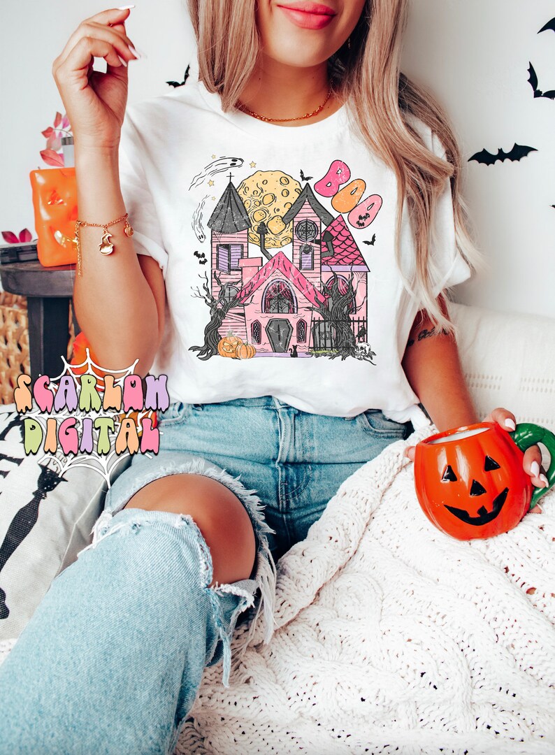 May include: A white t-shirt with a pink and white illustration of a haunted house with a full moon and bats. The text "BOO" is above the house. The model is wearing the shirt and blue jeans.