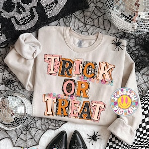 May include: A beige sweatshirt with the words "TRICK OR TREAT" in a colorful, block letter design. The letters have orange, black, and pink accents. The sweatshirt is part of a Halloween-themed collection.