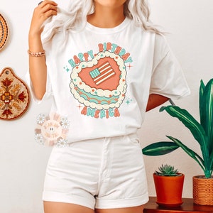 K&ouml;nnte beinhalten: Wei&szlig;es T-Shirt mit einem Grafikdruck eines herzf&ouml;rmigen Kuchens mit einer amerikanischen Flagge darauf. Der Text "Happy Birthday America" ist um den Kuchen herum geschrieben.