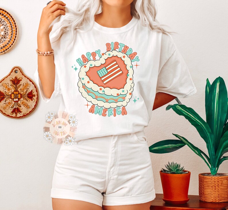 K&ouml;nnte beinhalten: Wei&szlig;es T-Shirt mit einem Grafikdruck eines herzf&ouml;rmigen Kuchens mit einer amerikanischen Flagge darauf. Der Text "Happy Birthday America" ist um den Kuchen herum geschrieben.