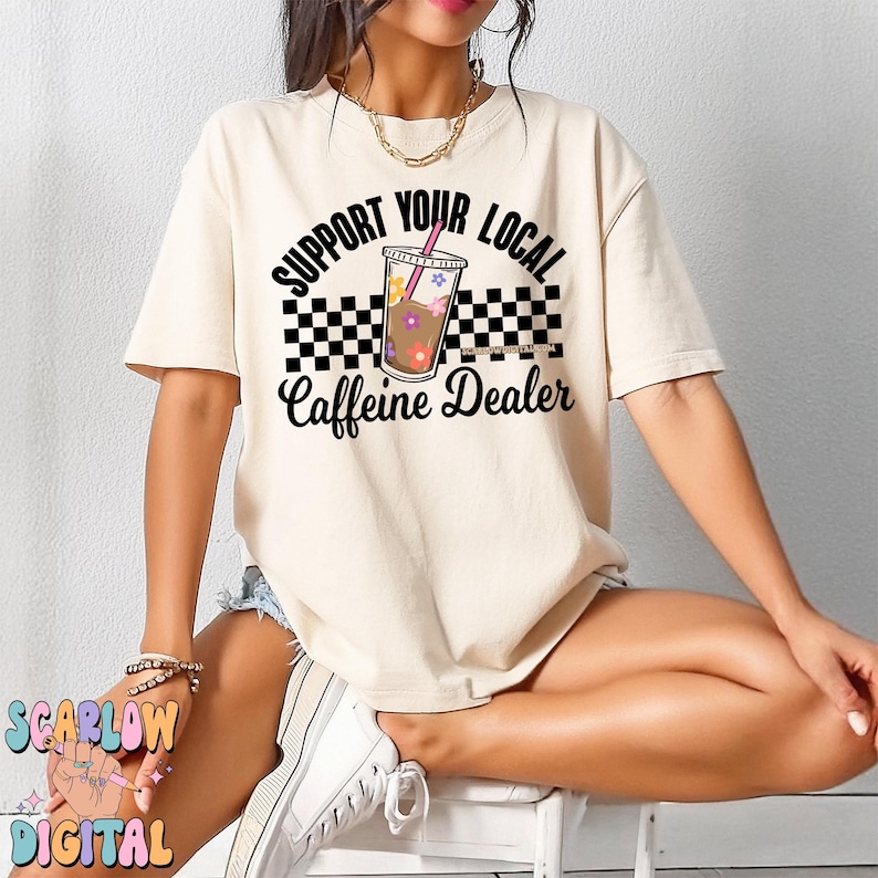 May include: A beige t-shirt with a black and white checkered design and the text "Support Your Local Caffeine Dealer" with a cartoon image of a cup of iced coffee with a pink straw and flowers.