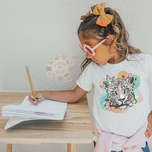 May include: A young person wearing pink sunglasses and a white t-shirt with a colorful graphic of a leopard's face surrounded by flowers and butterflies. They are sitting at a wooden table and writing in a notebook with a pencil.