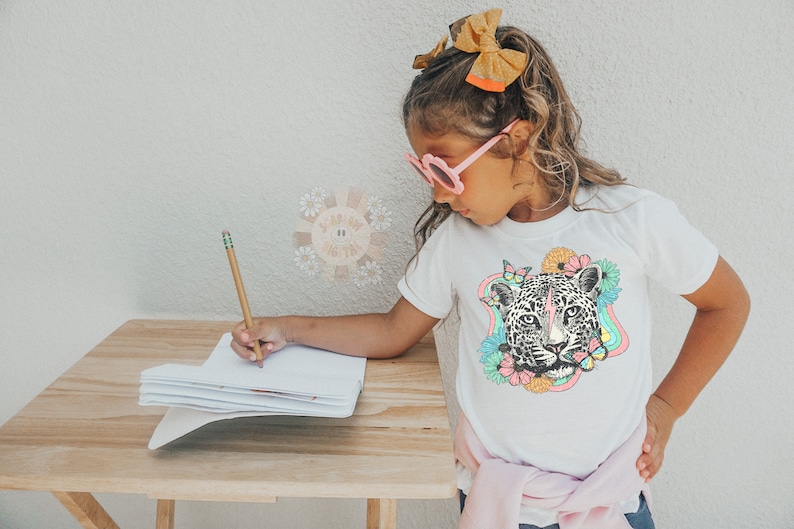 May include: A young person wearing pink sunglasses and a white t-shirt with a colorful graphic of a leopard's face surrounded by flowers and butterflies. They are sitting at a wooden table and writing in a notebook with a pencil.