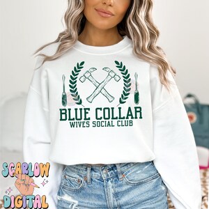 May include: White sweatshirt with a green graphic design of two crossed hammers, a wreath of leaves, and two screwdrivers. The text "Blue Collar Wives Social Club" is printed below the graphic.