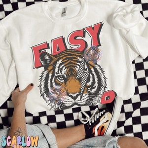 Puede incluir: Sudadera blanca con un gr&aacute;fico de tigre y la palabra "EASY" en letras rojas. El tigre tiene un rayo de color blanco y negro a cuadros sobre su ojo.