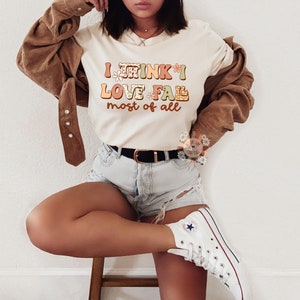 Op de afbeelding: Een wit T-shirt met de tekst "I think I love fall most of all" in kleurrijke, gestileerde letters met bloemmotieven.